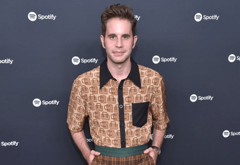 actor ben platt standing in front of a blue background with the spotify logo scattered over it
