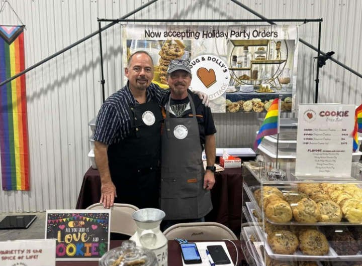 Two people standing behind a cookie booth at a market, with cookies on display and a sign about holiday party orders. Rainbow flags are visible in the background.