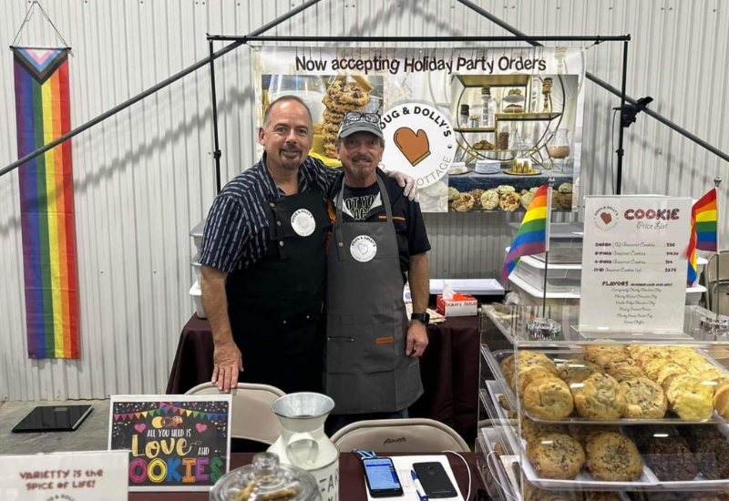Two people standing behind a cookie booth at a market, with cookies on display and a sign about holiday party orders. Rainbow flags are visible in the background.