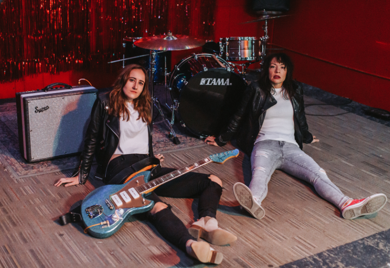 Two people sitting on stage with a guitar, amp, and drum set in the background.