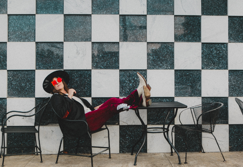 Person relaxing on a chair with feet on a table, against a checkered wall, wearing a hat with flowers.