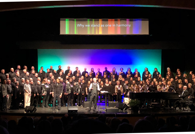 Choir performing on stage with conductor in front, text "Why we stand as one in harmony" above.