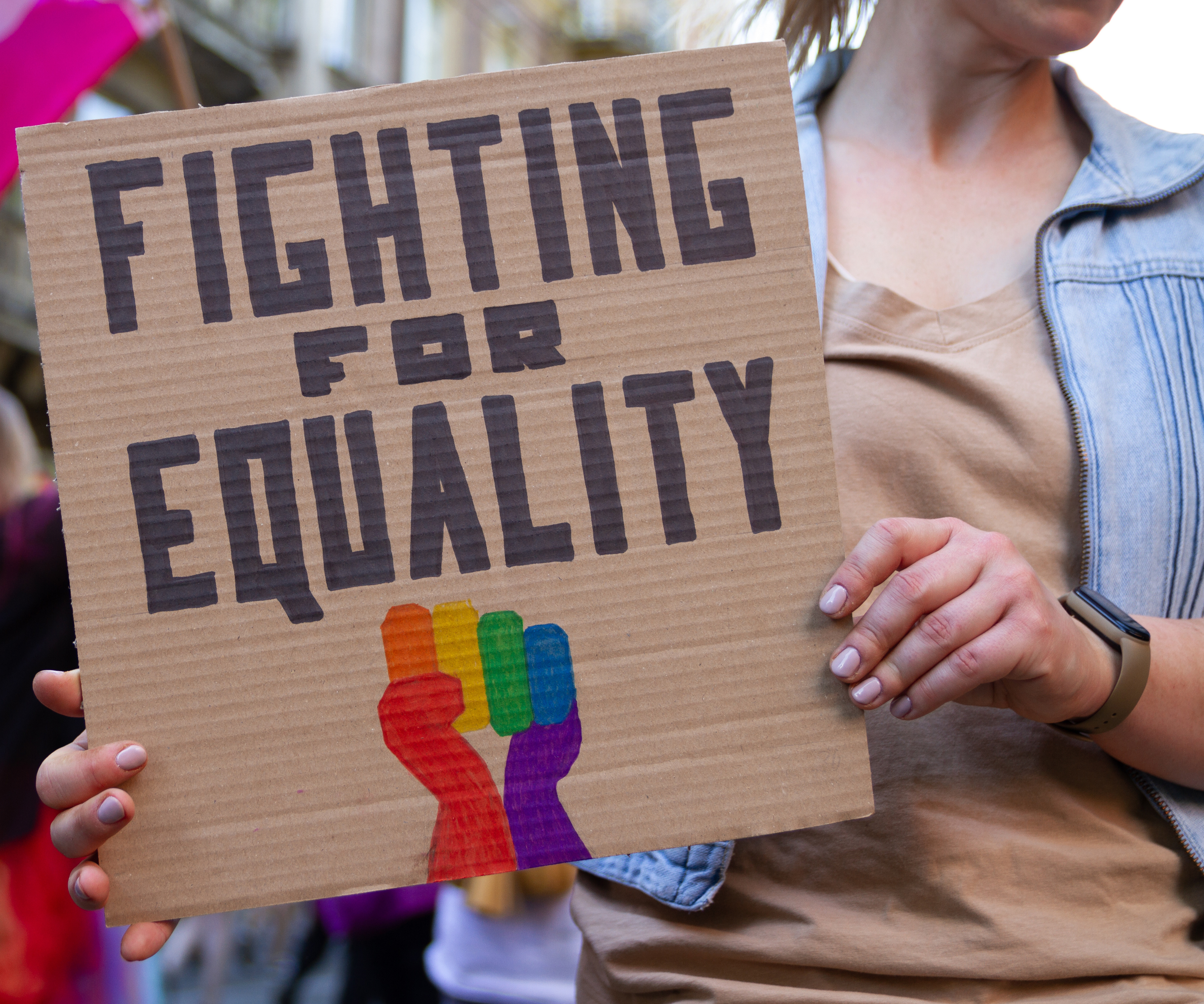 Person holding a sign that reads "FIGHTING FOR EQUALITY" with a rainbow-colored fist.