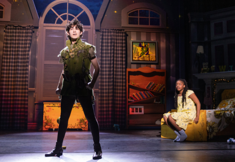 A stage scene with two actors, colorful set design resembling a child's bedroom, and ambient lighting.