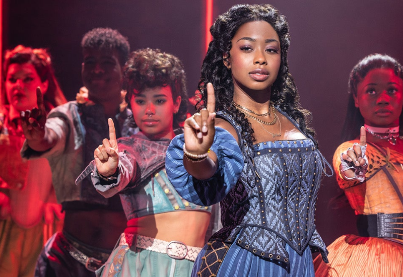A group of performers in vibrant costumes stands under dramatic lighting, showcasing intricate designs and raising their hands in unison.