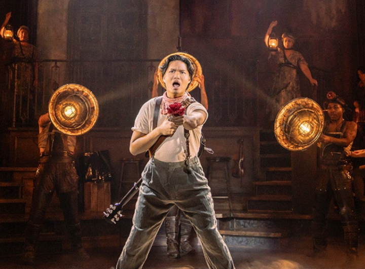 A performer in overalls holds a flower and strikes a dynamic pose on stage, surrounded by others with vintage lanterns and theatrical attire.