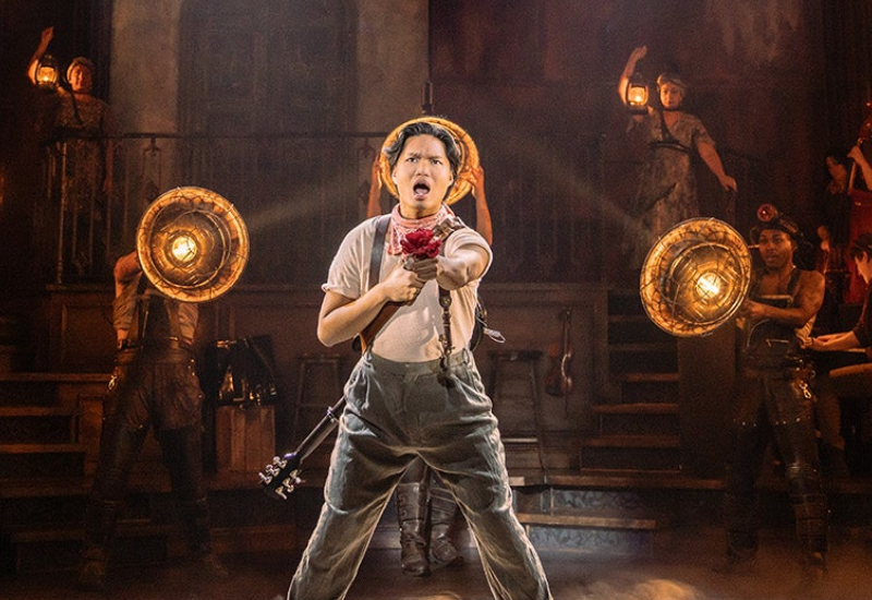 A performer in overalls holds a flower and strikes a dynamic pose on stage, surrounded by others with vintage lanterns and theatrical attire.