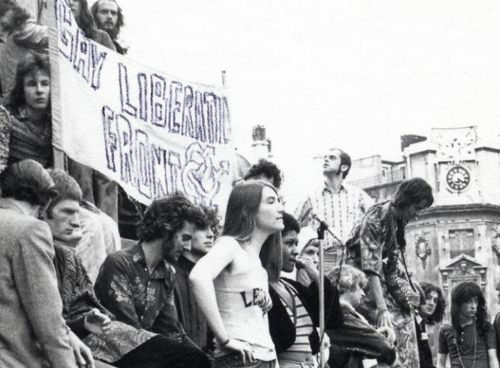 A group of activists gathering outdoors, holding a banner that reads "Gay Liberation Front," with historic buildings in the background.