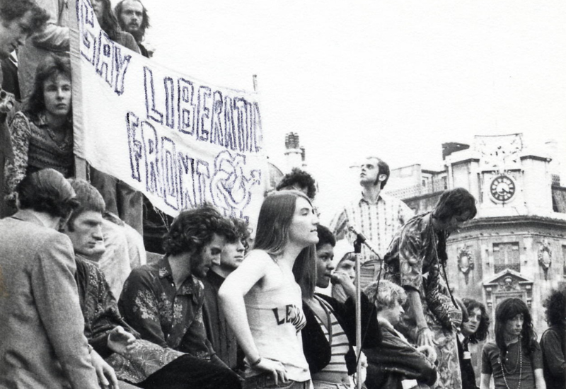 A group of activists gathering outdoors, holding a banner that reads "Gay Liberation Front," with historic buildings in the background.