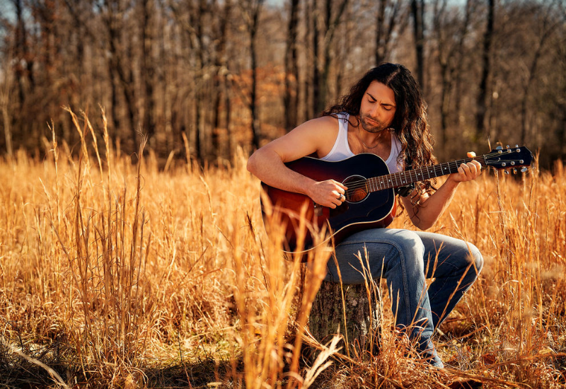 A person sits on a log in a golden field, playing an acoustic guitar surrounded by tall grass and trees in the background.