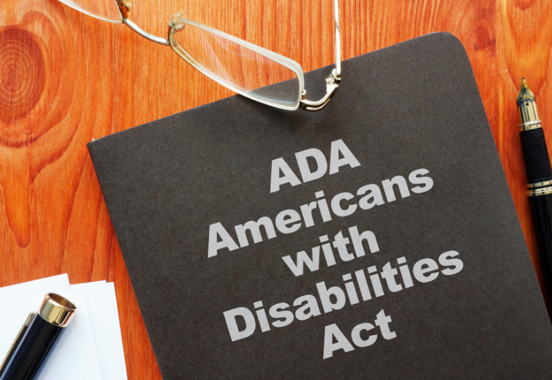 A black notebook titled "ADA Americans with Disabilities Act" lies on a wooden table, accompanied by reading glasses and pens.
