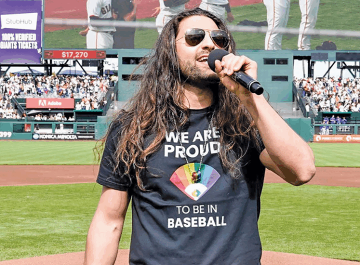 A performer in a black t-shirt with a rainbow design stands on a baseball field, microphone in hand, addressing a cheering crowd.