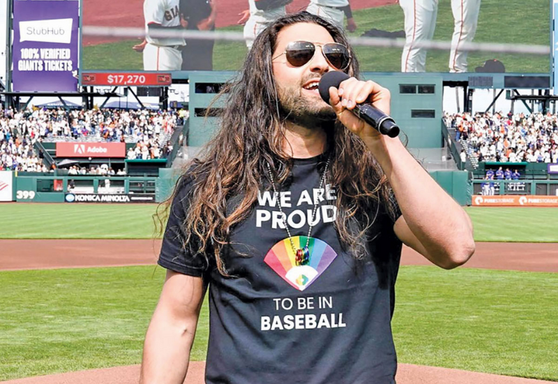 A performer in a black t-shirt with a rainbow design stands on a baseball field, microphone in hand, addressing a cheering crowd.
