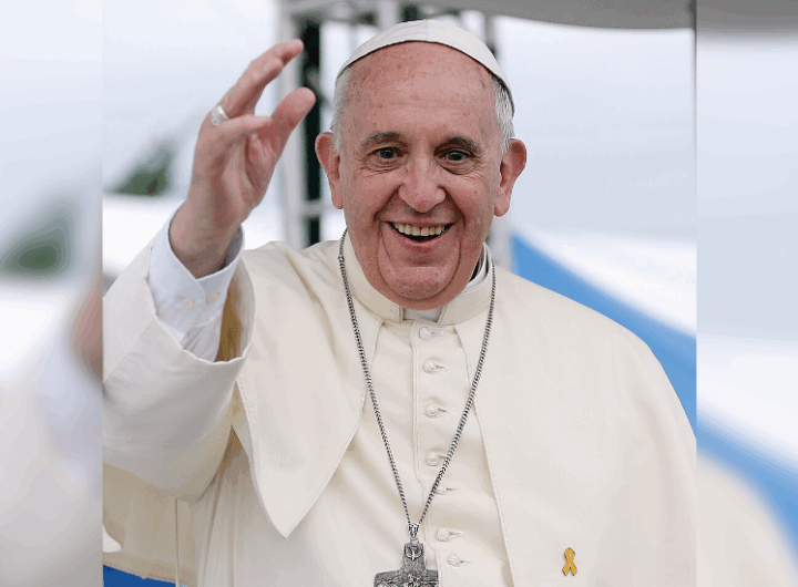 The Pope, dressed in white robes, raises his right hand in greeting against a blurred outdoor background. A yellow ribbon adorns his attire.