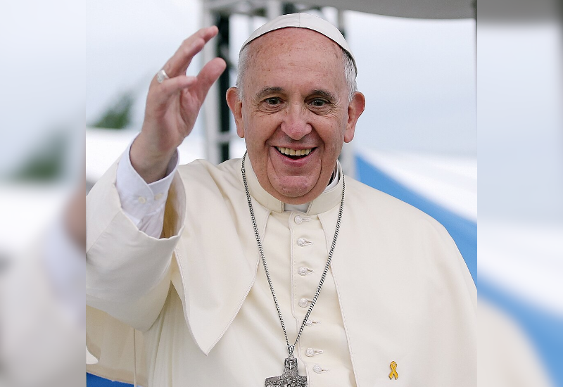 The Pope, dressed in white robes, raises his right hand in greeting against a blurred outdoor background. A yellow ribbon adorns his attire.
