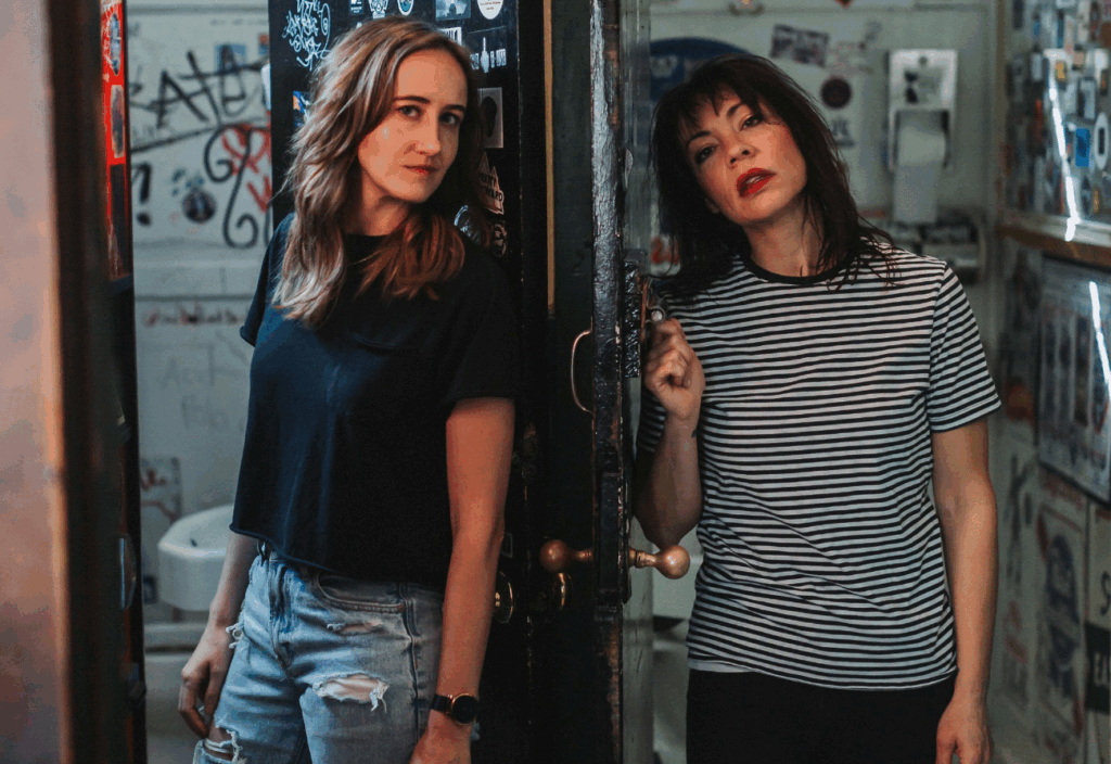 Two women pose near a partially open door, showcasing a vibrant, graffiti-covered interior with colorful stickers on the walls.
