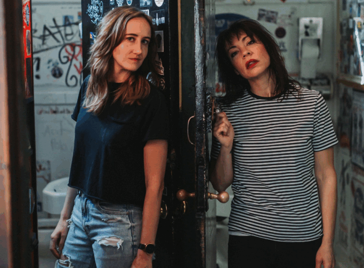 Two women pose near a partially open door, showcasing a vibrant, graffiti-covered interior with colorful stickers on the walls.