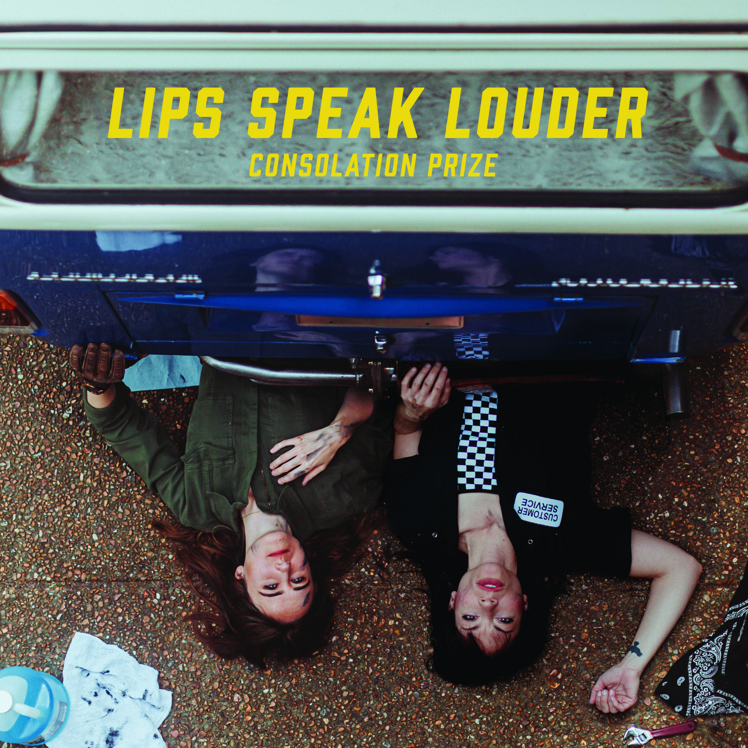 Two individuals lie on the ground beside a vintage blue vehicle, surrounded by scattered items, with the title "Lips Speak Louder."