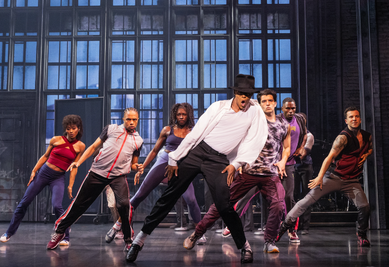 A group of diverse dancers in stylish outfits perform energetically on a brightly lit stage with large windows in the background.