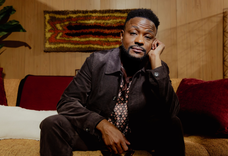 Wyn Starks, dressed in a stylish outfit, sits pensively on a couch surrounded by rich textiles and warm, earthy tones in a cozy room.