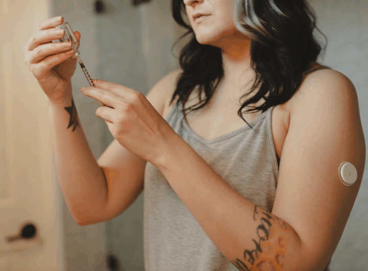 A woman in a tank top prepares a syringe, showcasing a wearable health device on her arm; she has visible arm tattoos.