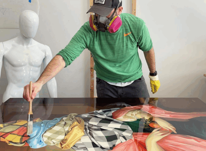 An artist in a green shirt and mask works on a colorful painting, using a brush over a detailed, vibrant surface. A white mannequin stands nearby.