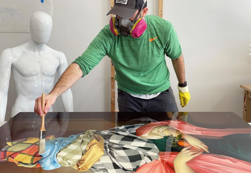 An artist in a green shirt and mask works on a colorful painting, using a brush over a detailed, vibrant surface. A white mannequin stands nearby.