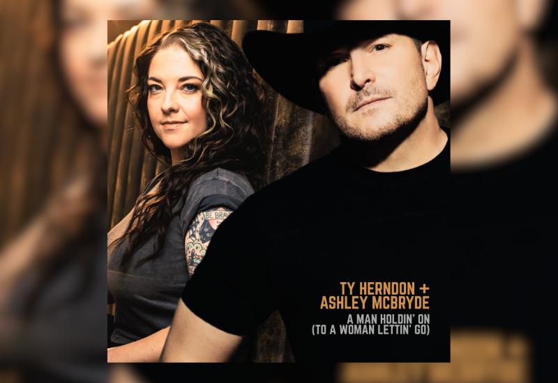 Ty Herndon and Ashley McBryde pose together in a dimly lit setting, showcasing their stylish outfits and shared musical connection.