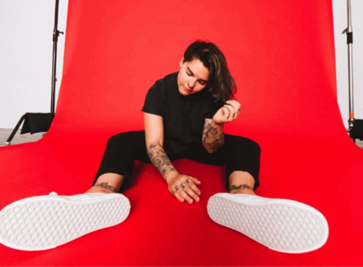 A person sits on a red background, wearing a black shirt and pants, with visible tattoos and white sneakers.