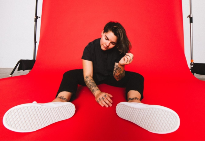 A person sits on a red background, wearing a black shirt and pants, with visible tattoos and white sneakers.