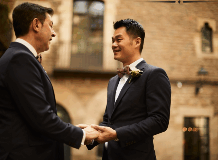 Two elegantly dressed men exchange vows while holding hands outdoors, surrounded by a rustic brick backdrop and warm lighting.