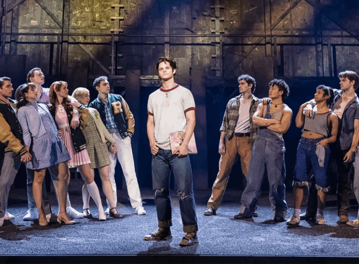 A group of diverse performers in colorful costumes poses dramatically on a theater stage, showcasing a lively musical scene.