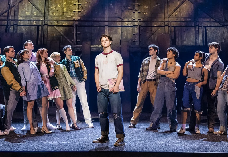 A group of diverse performers in colorful costumes poses dramatically on a theater stage, showcasing a lively musical scene.