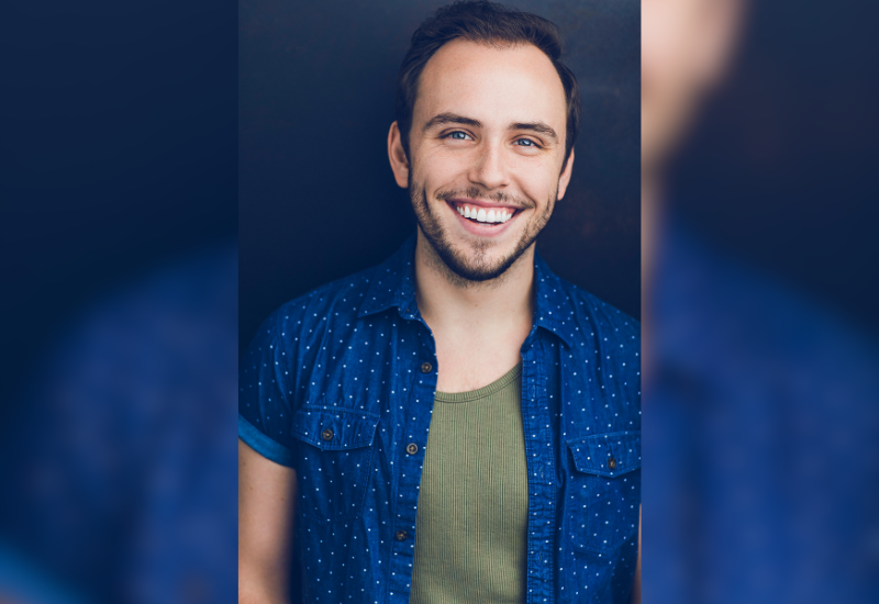 A person wearing a blue polka dot shirt over a green tank top stands against a dark background, with arms relaxed at their sides.