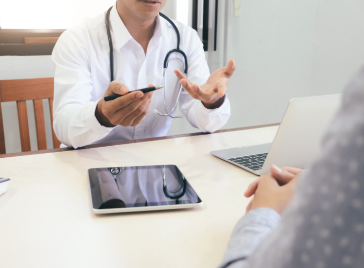A doctor explains treatment options to a patient during a consultation, with a laptop and tablet on the table.