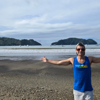 A person stands on a sandy beach with arms outstretched, overlooking a calm sea and distant green hills under a partly cloudy sky.