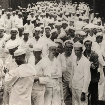 Black and white photo of a large group of sailors wearing white uniforms, gathered closely together, outdoors near a building.