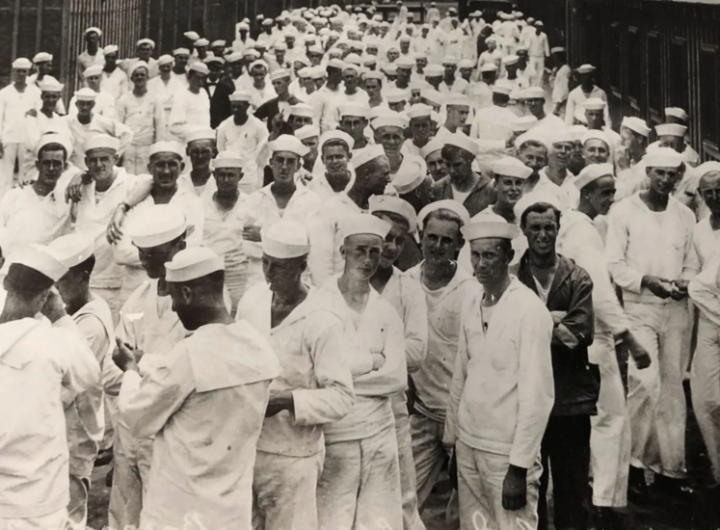 Black and white photo of a large group of sailors wearing white uniforms, gathered closely together, outdoors near a building.
