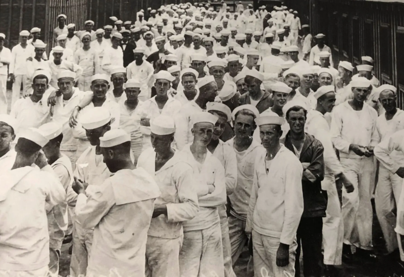 Black and white photo of a large group of sailors wearing white uniforms, gathered closely together, outdoors near a building.