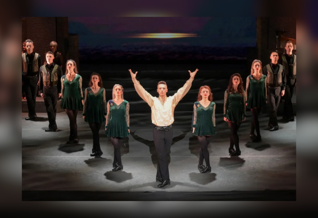 Dancers in green dresses and black pants performing an Irish step dance on stage with a sunset backdrop.