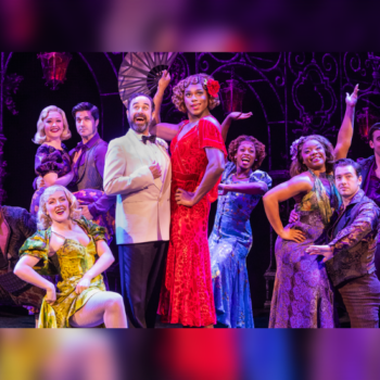 Theatrical cast dressed in vibrant 1920s-style costumes posed on stage with dramatic lighting and ornate backdrop.
