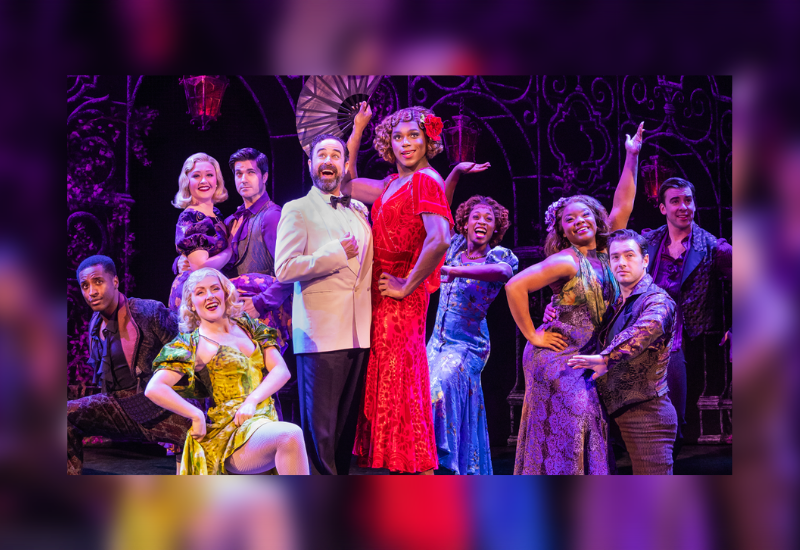 Theatrical cast dressed in vibrant 1920s-style costumes posed on stage with dramatic lighting and ornate backdrop.