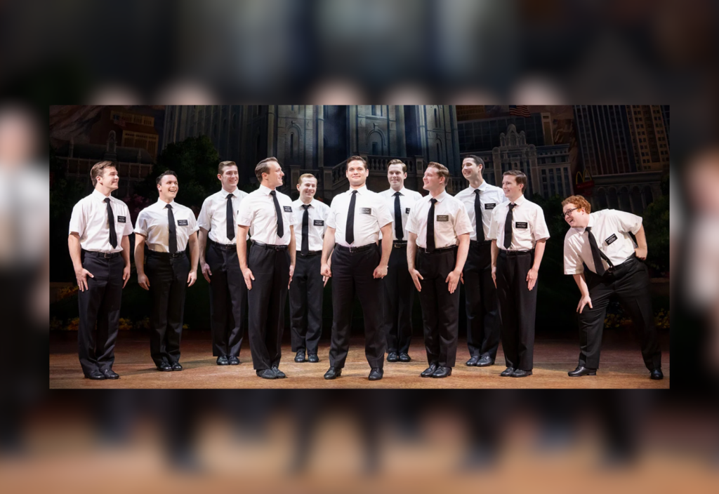 Group of men in white shirts, black ties, and black pants performing on stage with a cityscape backdrop.