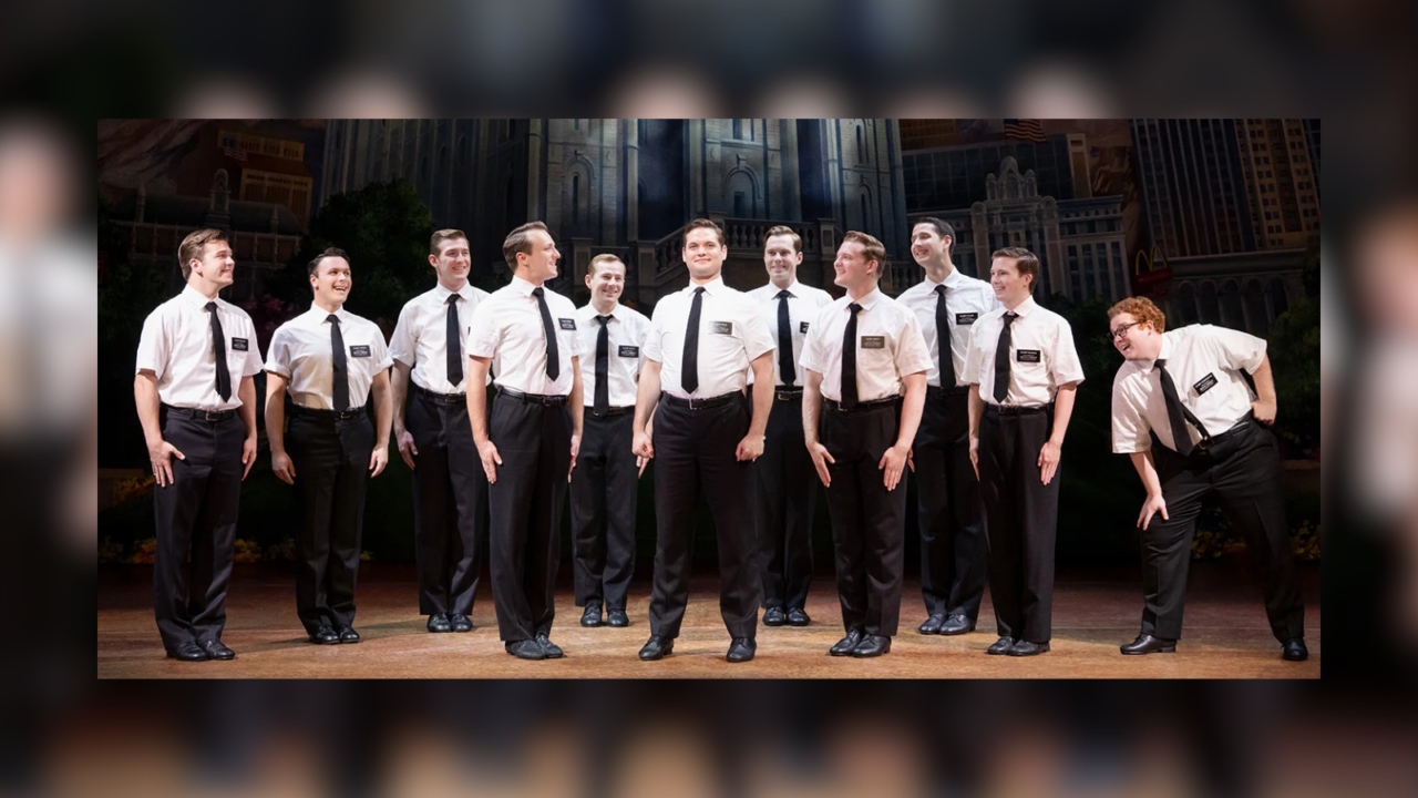 Group of men in white shirts, black ties, and black pants performing on stage with a cityscape backdrop.